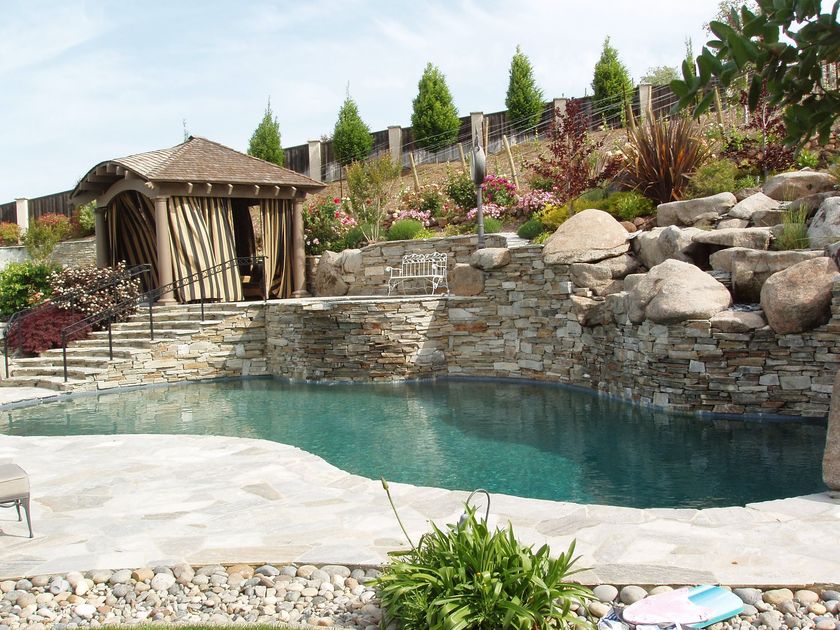 Backyard pool with stone waterfall, wooden gazebo, and landscaped rocks and flowers