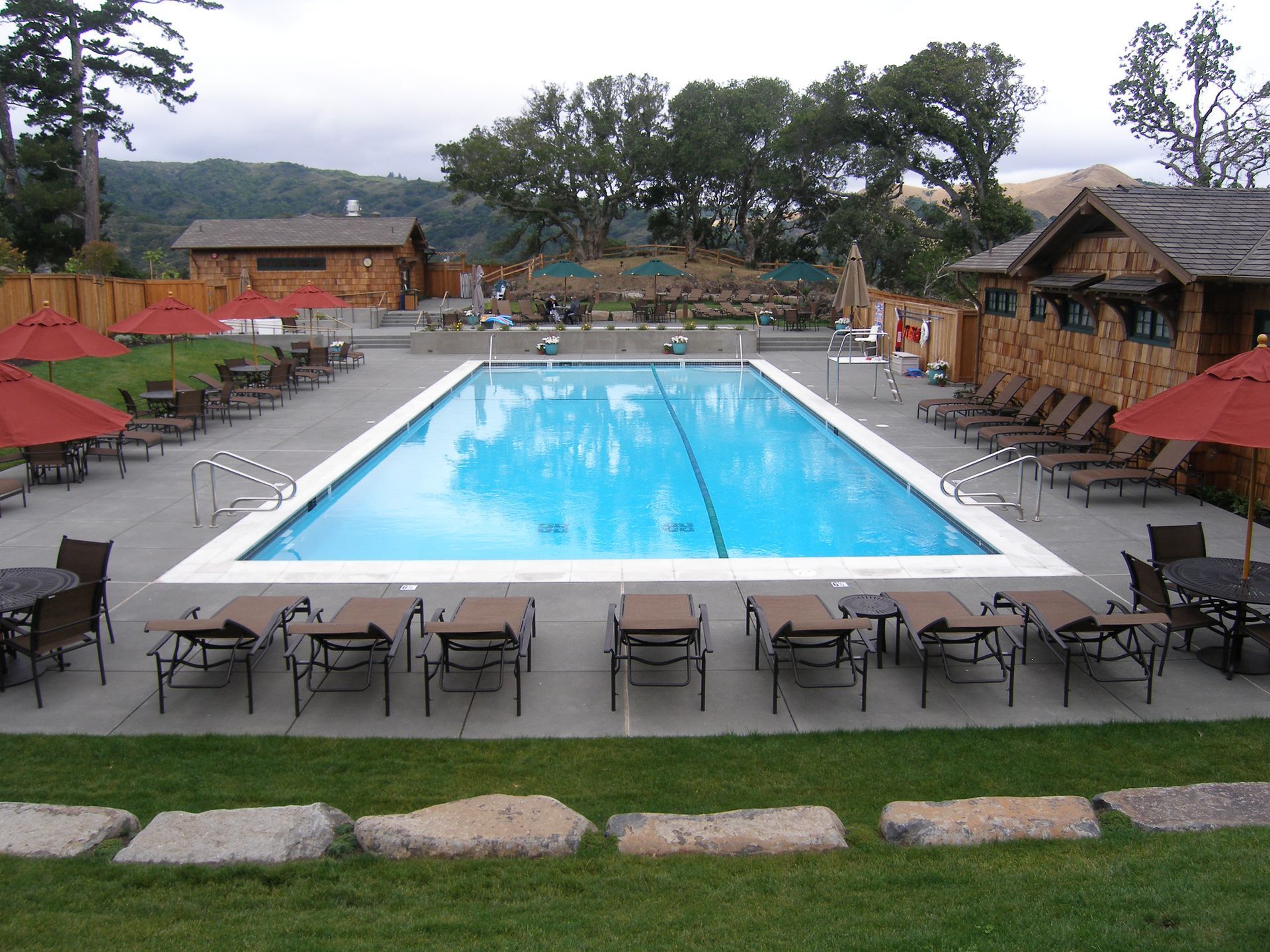 Outdoor pool with lounge chairs and tables, surrounded by thatched-roof cabanas and greenery