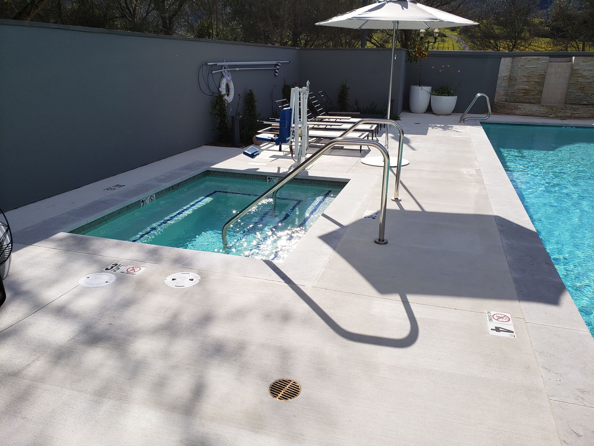 Backyard pool deck with a small raised hot tub, lounge chairs, umbrella, and bright sunlight