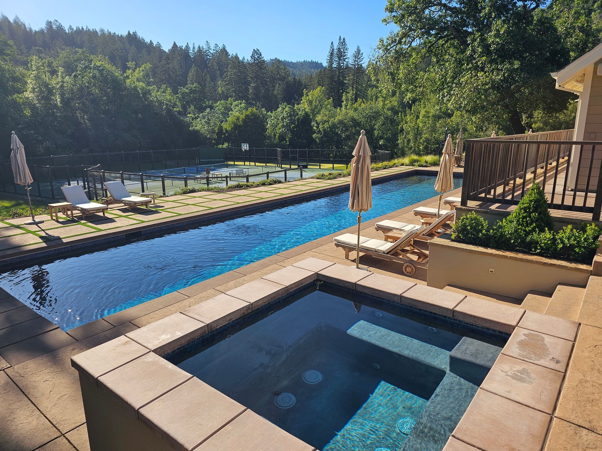 Backyard pool and hot tub with lounge chairs beside a sunny deck and wooded view