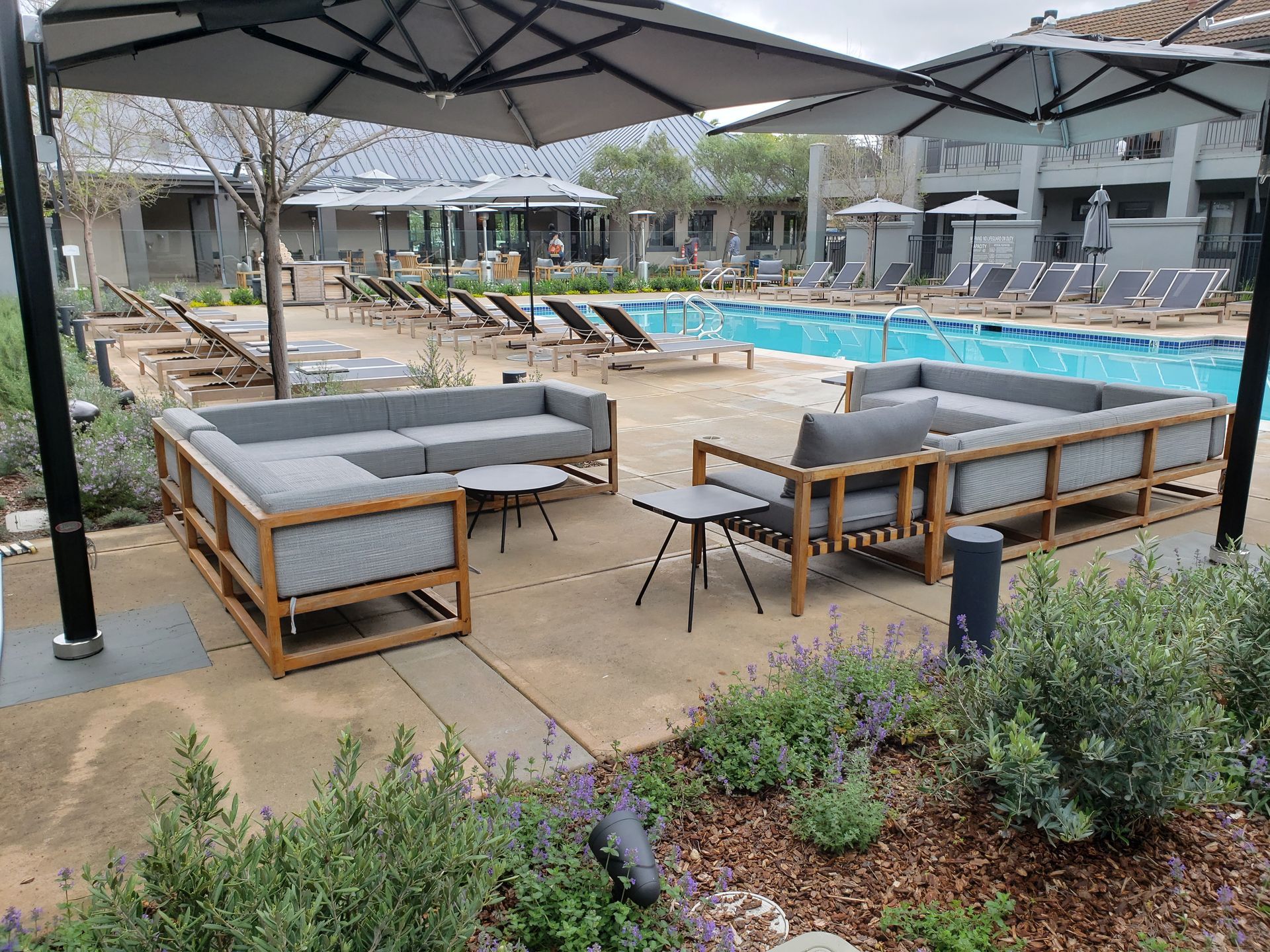 Outdoor pool area with gray lounge seating, umbrellas, and a wooden deck beside a blue swimming pool