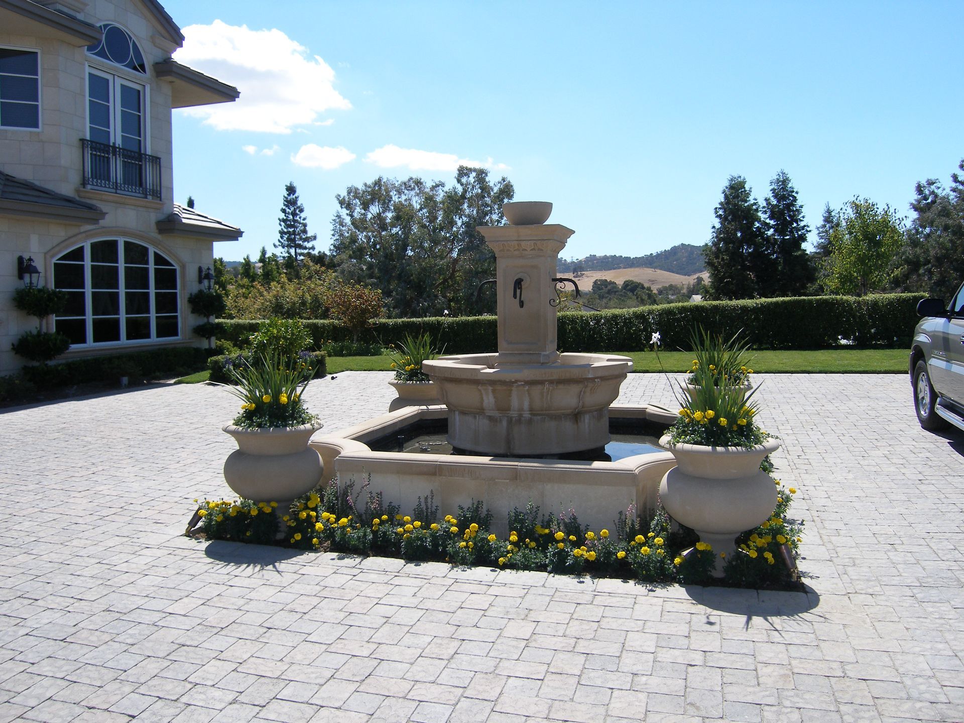 Stone fountain in a landscaped driveway courtyard beside a house, with potted flowers and parked car.