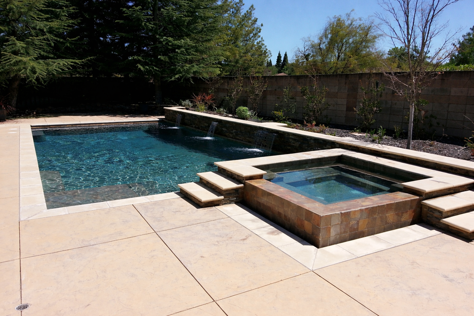 Backyard pool with raised spa and beige patio, surrounded by trees and a fence.