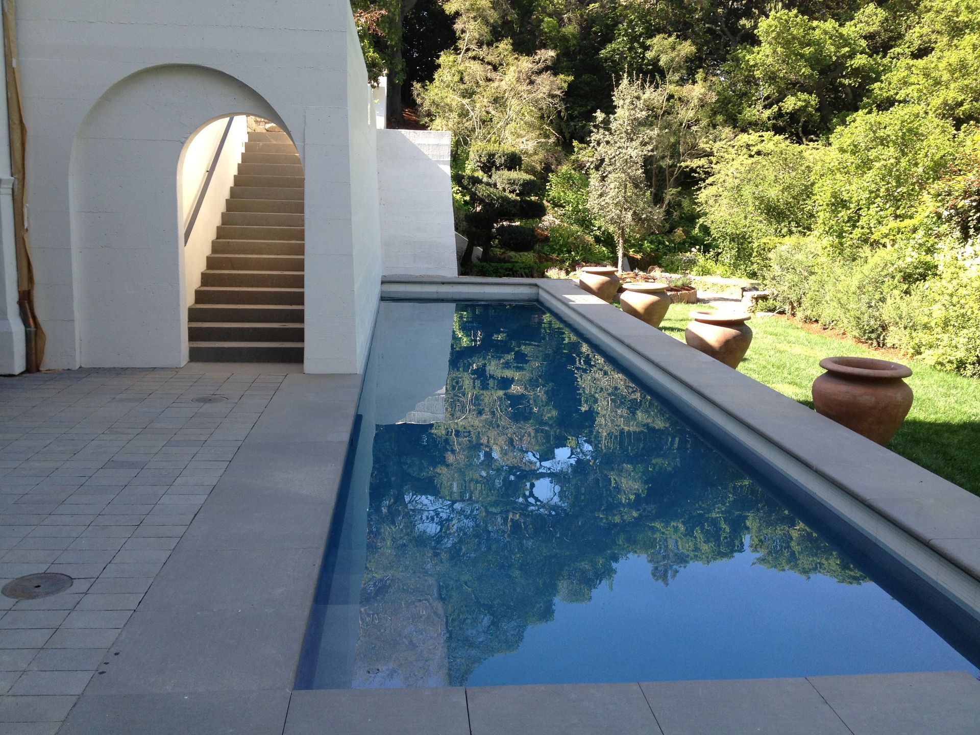 Courtyard pool beside white archway and stairs, bordered by greenery and stone pots