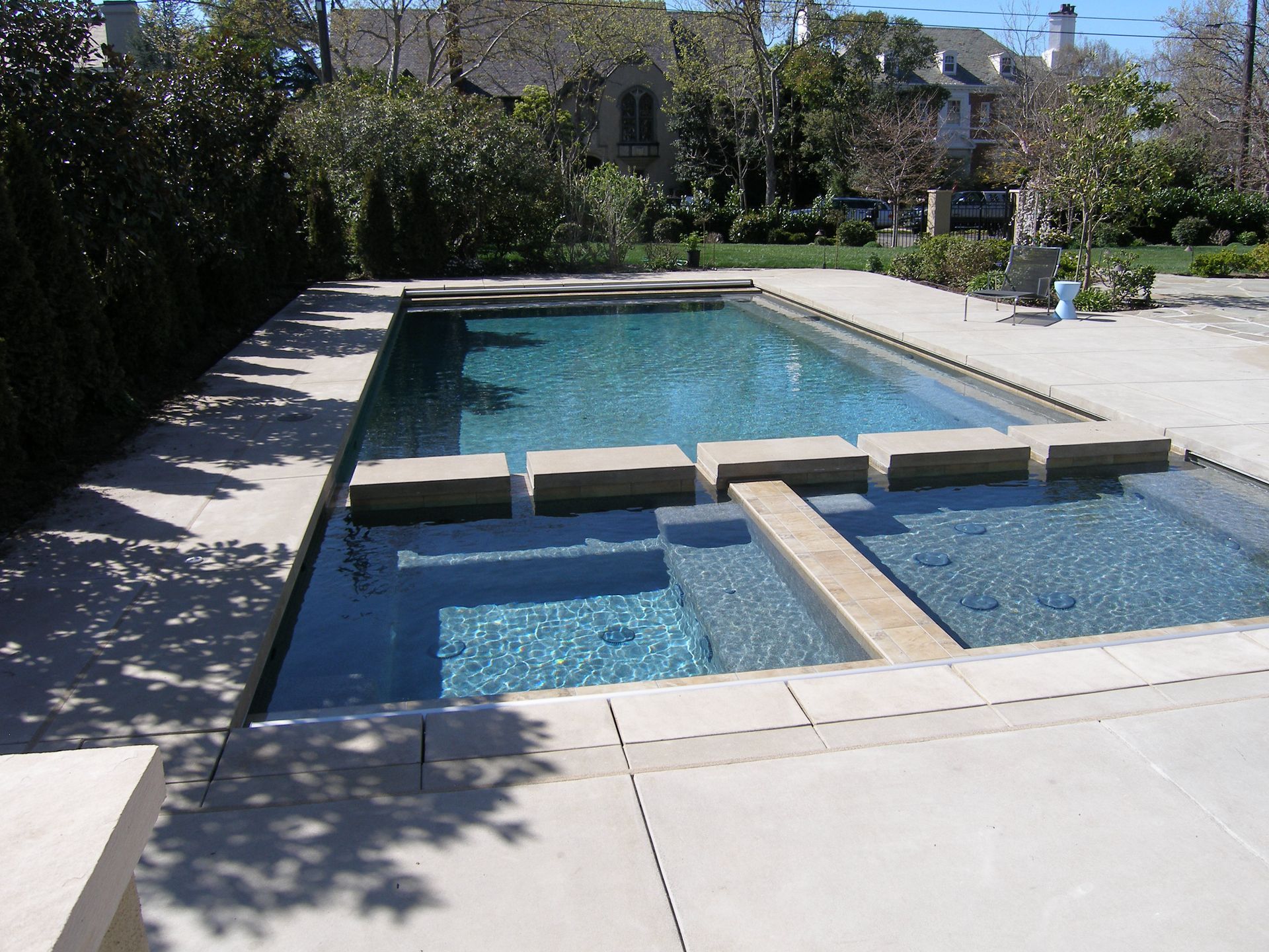 Modern backyard swimming pool with a raised spa and stone patio, surrounded by trees and houses