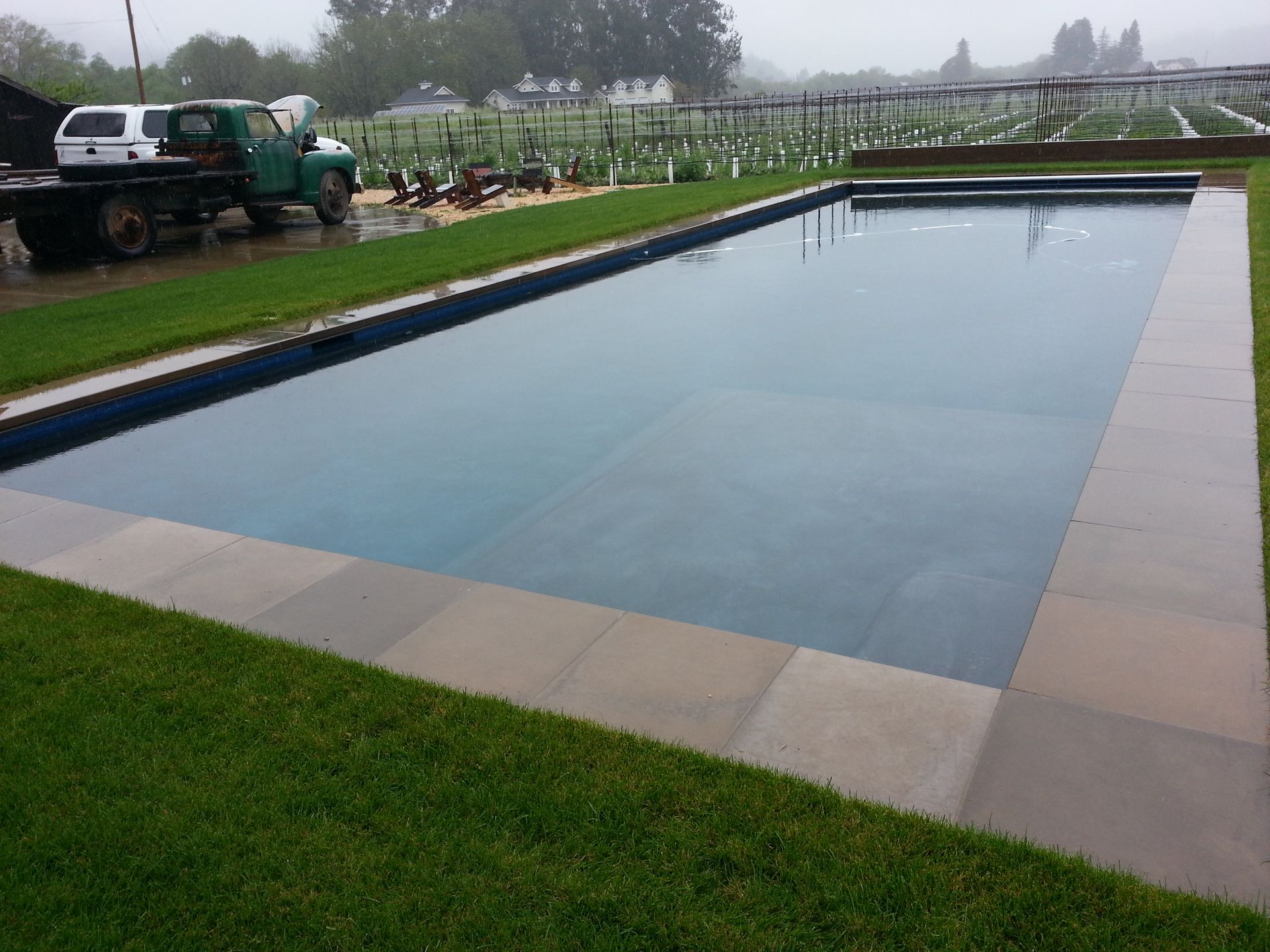 Rectangular outdoor swimming pool with still water, bordered by grass and paved edges, on a cloudy day