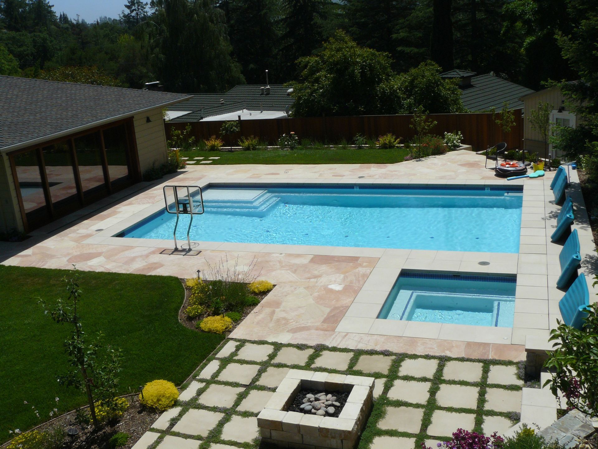 Backyard pool with hot tub, lounge chairs, and paved patio surrounded by greenery