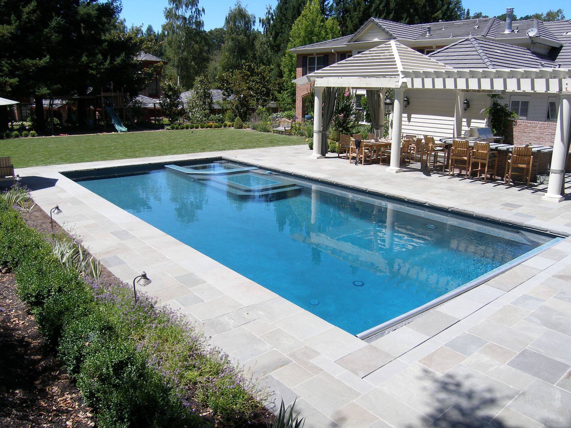 Rectangular backyard swimming pool beside a white pergola and patio chairs on a sunny day