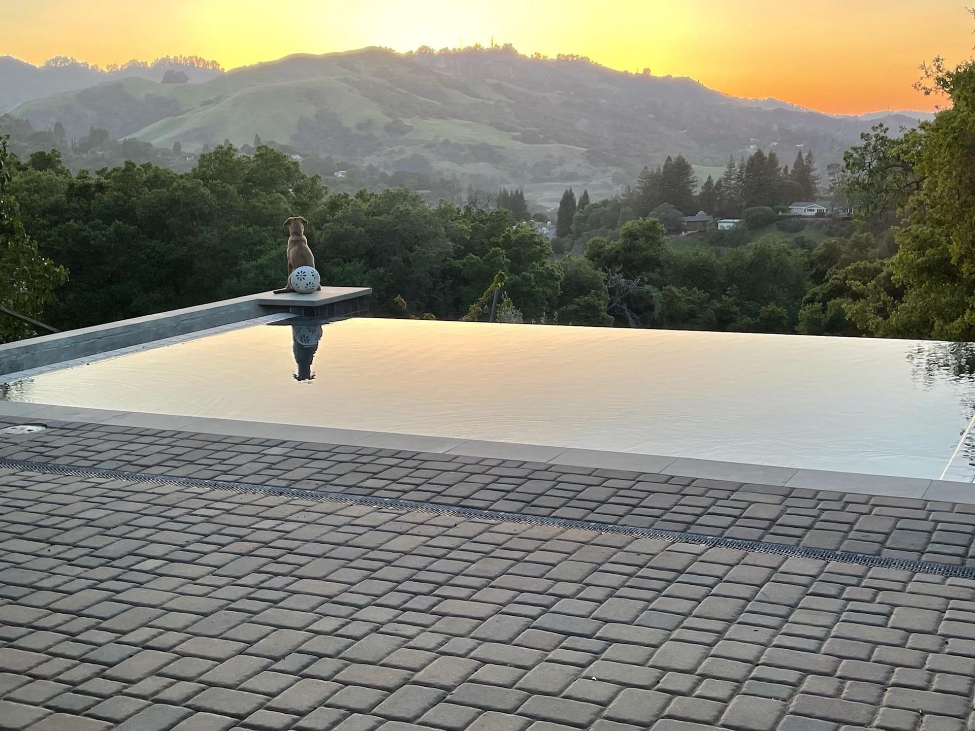 Infinity pool at sunset reflecting golden sky, with a person meditating and distant green hills.