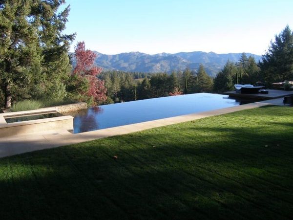 Modern house with an infinity pool overlooking a tree line. Two lounge chairs, a striped umbrella, and blue sky.