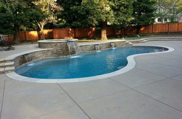 Swimming pool with waterfalls and stone accents surrounded by a concrete patio, fence, and trees.