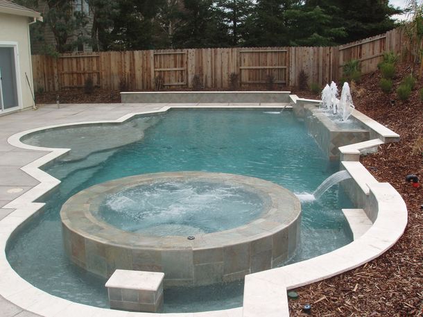 Swimming pool with attached jacuzzi and fountains, surrounded by concrete and a wooden fence.