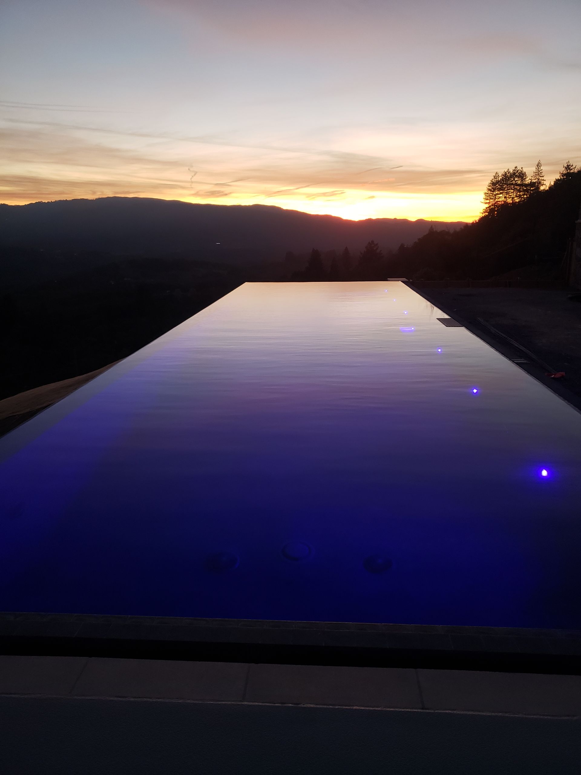 Sunset over a long reflective pool with blue lights along the edge, framed by dark hills.