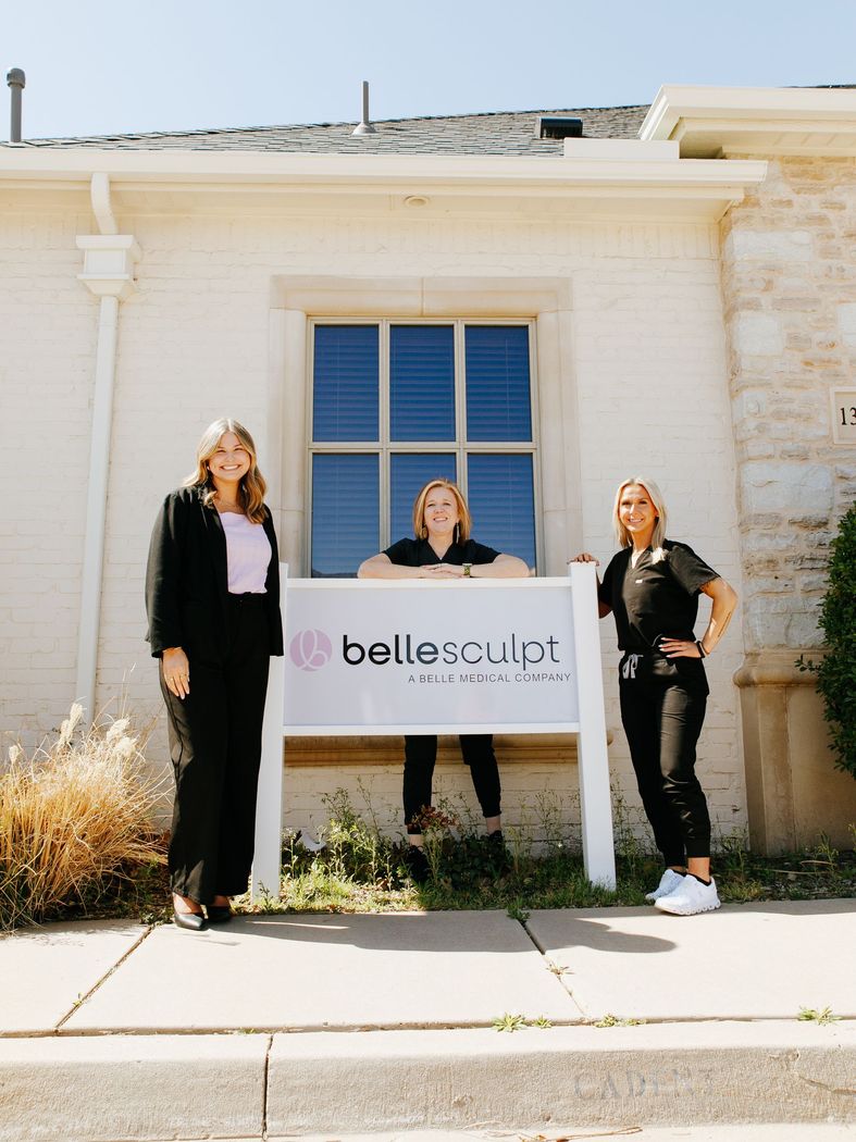 Three people standing outside a white building holding a “bellesculpt oklahoma” sign