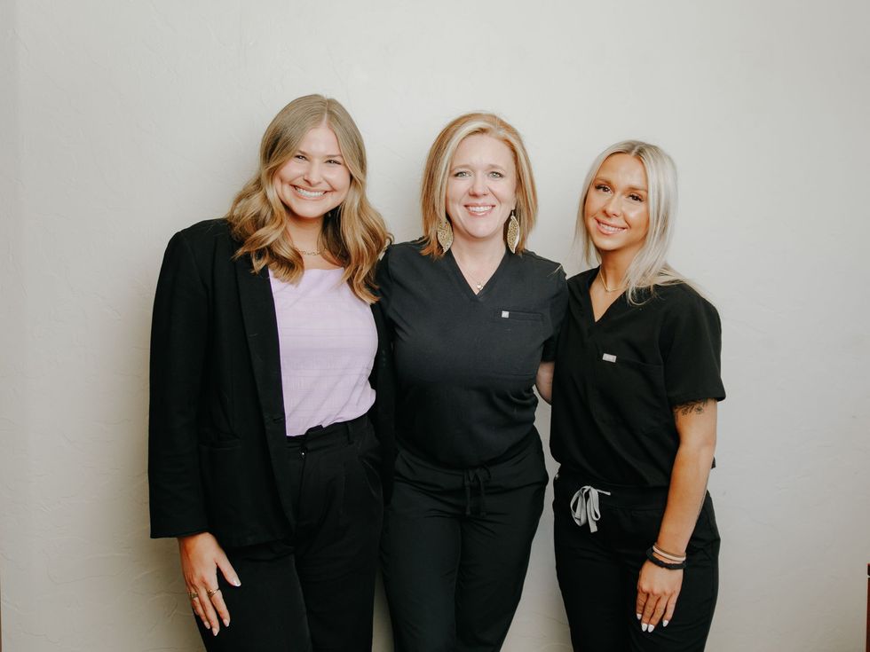 Three smiling clinic women pose together against a light wall, wearing black and light purple outfits.