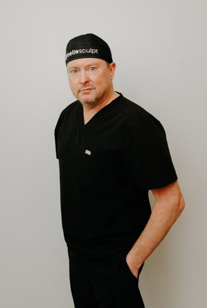 Doctor Blatchford in black shirt and cap standing against a light gray wall, one hand in his pocket