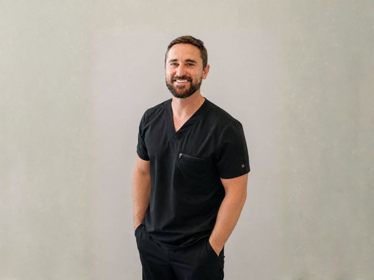 Smiling male doctor in black scrubs standing against a light wall with hands in pockets