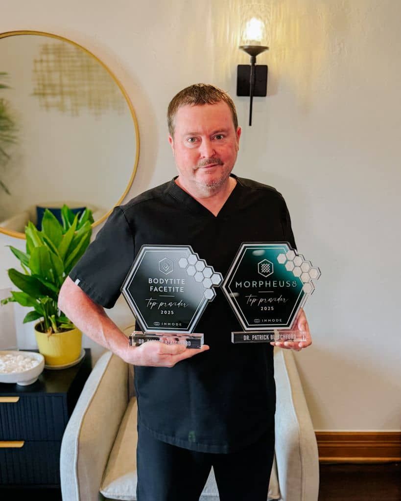National Top Provider Awards Doctor Blatchford holding two clear awards, standing indoors beside a plant and wall sconce.