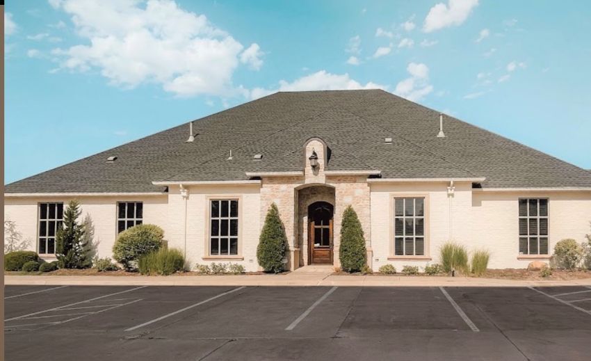 Single-story beige building with a dark roof, arched entrance, and parking lot under a blue sky