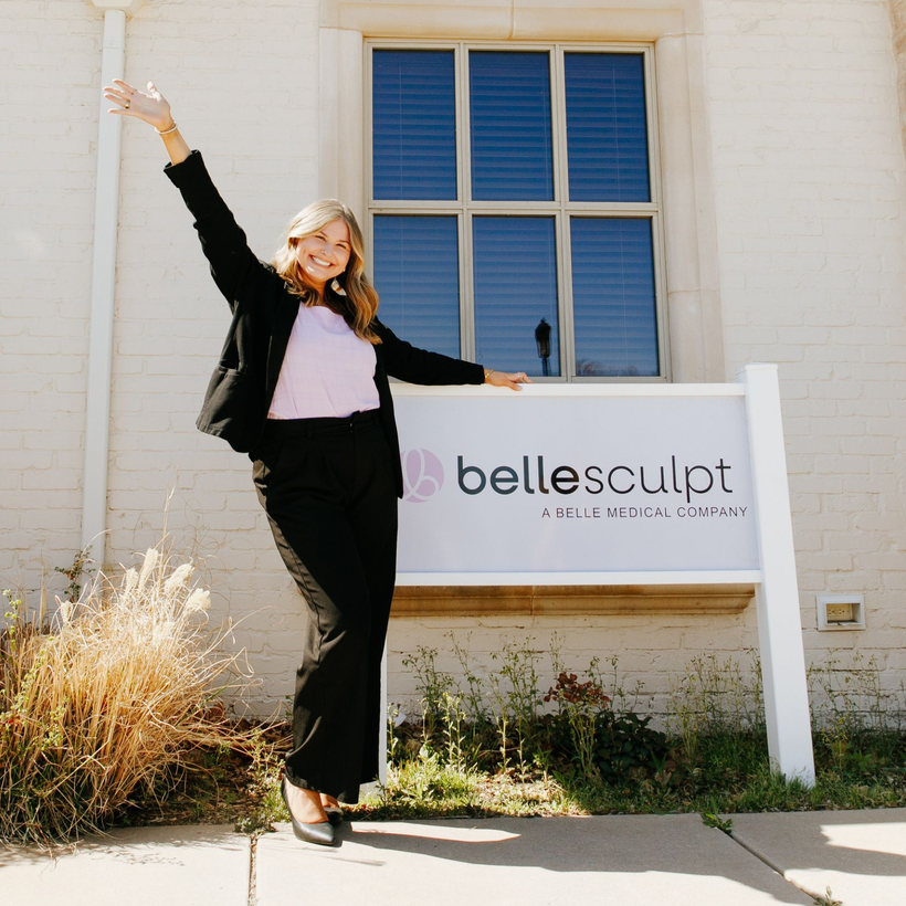 Woman in black posing beside a Bellesculpt sign outside a white building