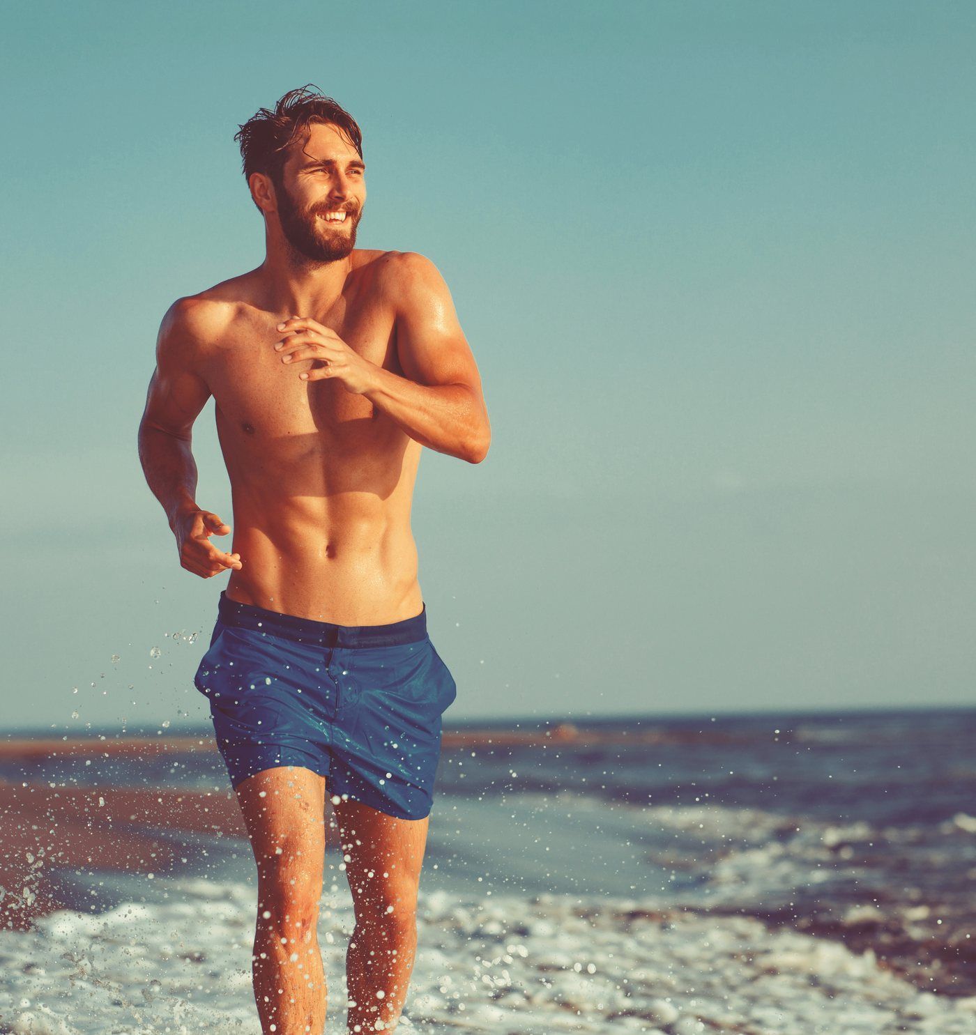 Fit, bearded man smiling as he jogs along the beach with a lean, defined midsection