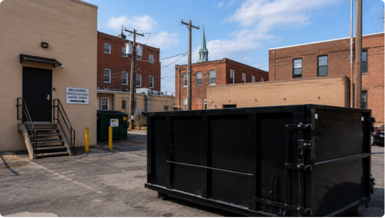 Back alley with a large black dumpster beside brick buildings and a small stairway loading entrance