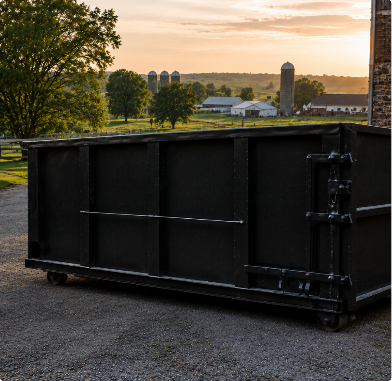 Large black dumpster on gravel at sunset with farm silos and trees in the background