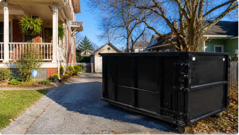 Large black dumpster in a gravel driveway beside a house on a sunny day