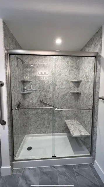 A walk-in shower with gray marbled walls, a white base, a bench, grab bars, and a sliding glass door.