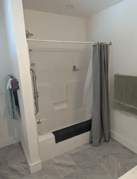A white bathtub and shower stall with a grab bar, a gray shower curtain, and a black bath mat in a bathroom.