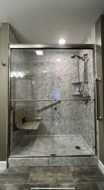 A walk-in shower with marble-look walls, a glass sliding door, a wall-mounted fold-down bench, and safety grab bars.