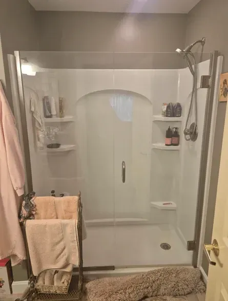 A glass-enclosed shower with white walls and built-in corner shelves, featuring a chrome shower head and a towel rack.