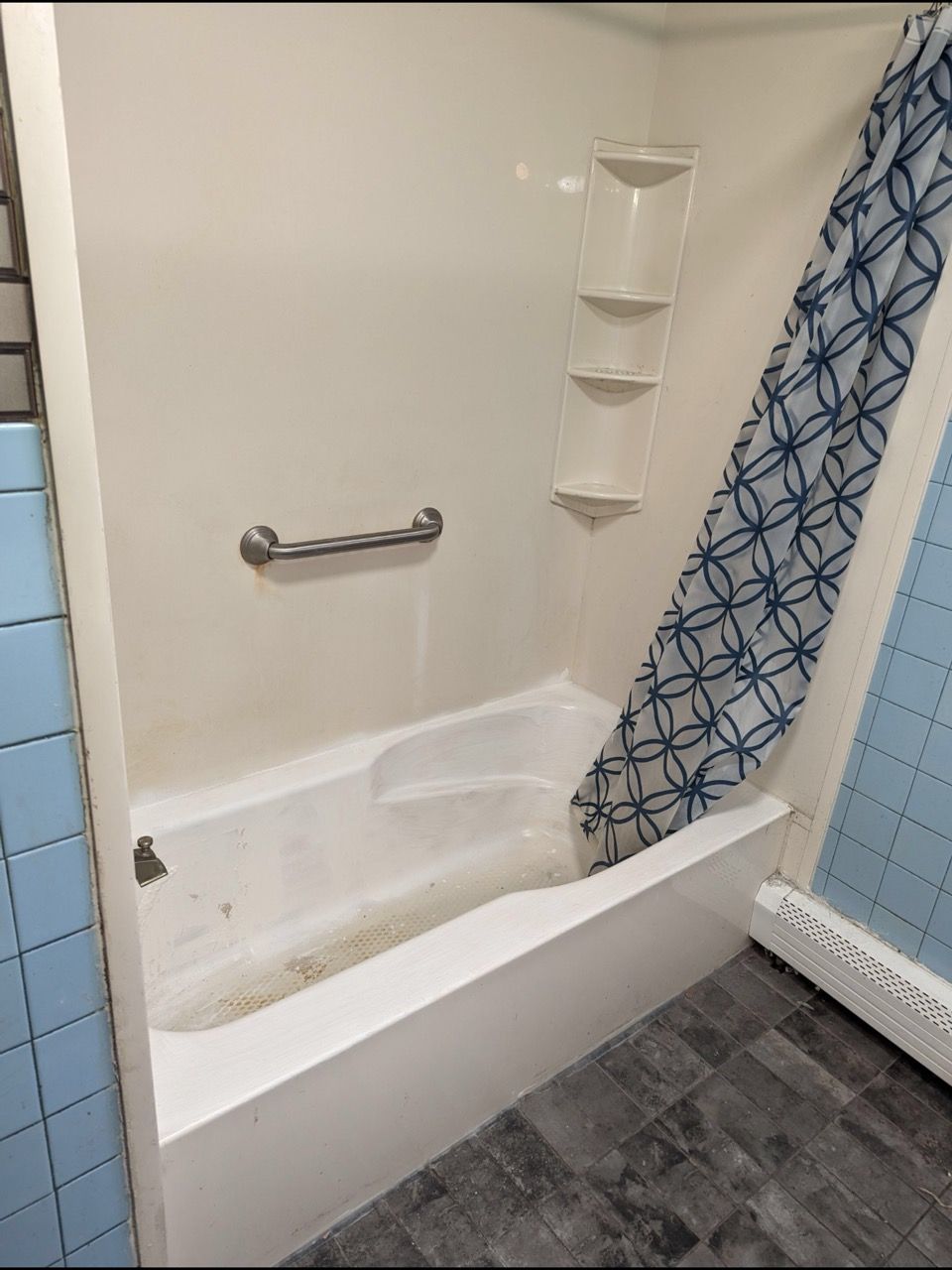 White bathtub with a metal grab bar on the back wall and a built-in shelving unit, next to a blue patterned shower curtain.