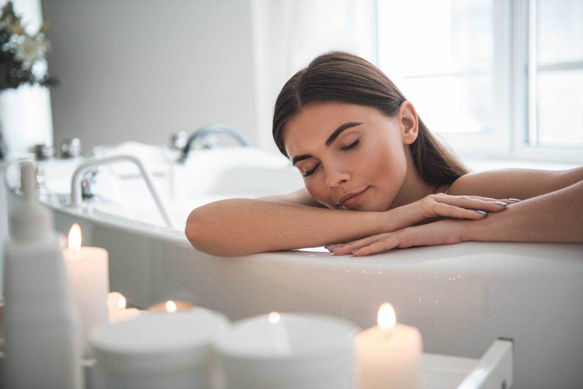 A person relaxing with eyes closed in a white bathtub, with candles in the foreground.