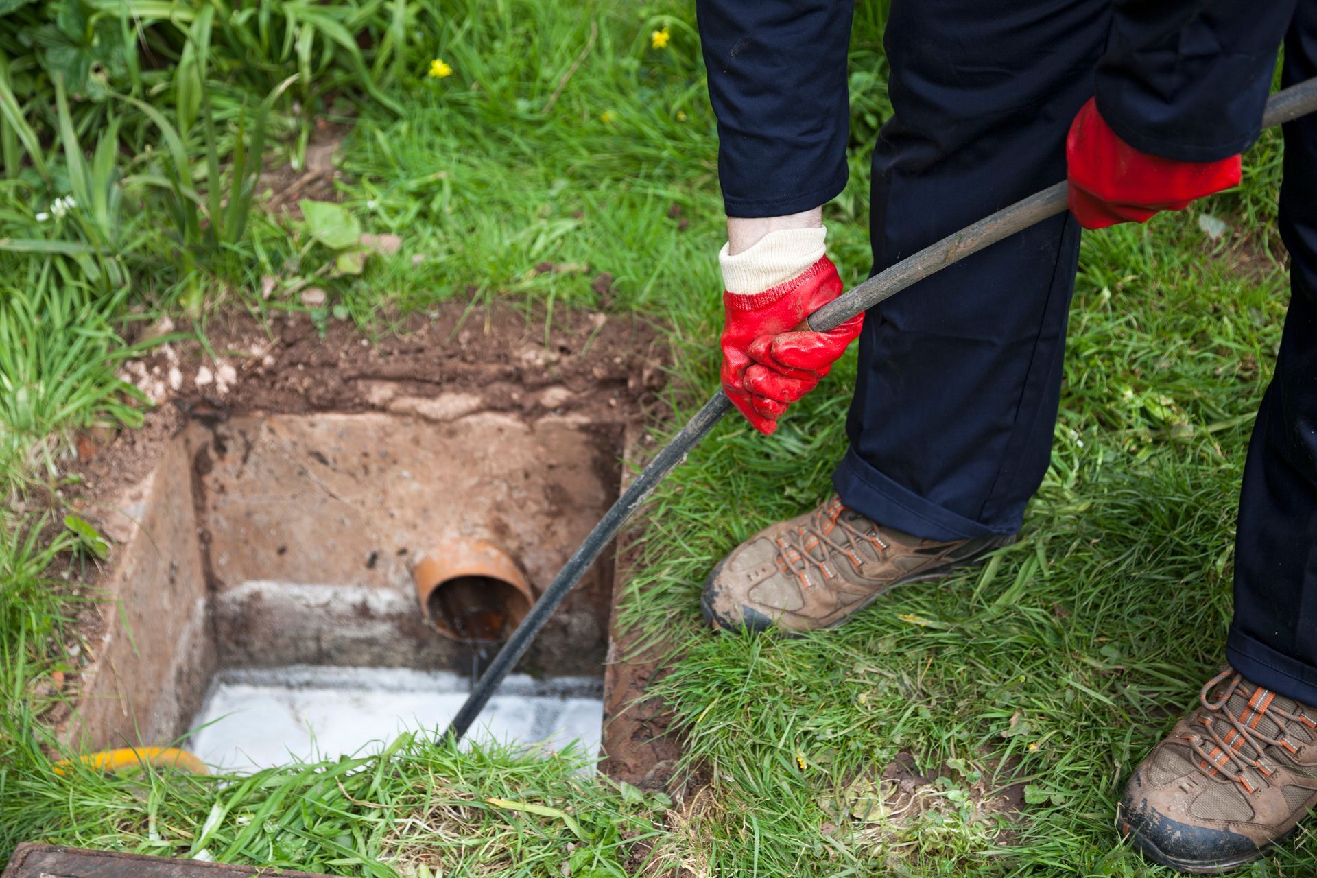 Worker in red gloves inspecting a muddy square pit with a pipe opening in the ground.