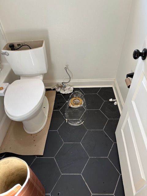 Small bathroom with a white toilet, black hex tile floor, and exposed toilet plumbing by the wall.