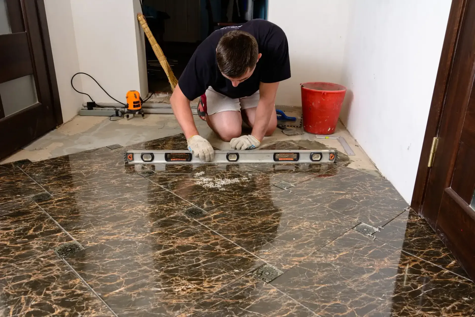 Man kneeling, using a level to install dark patterned floor tiles indoors, near a door.