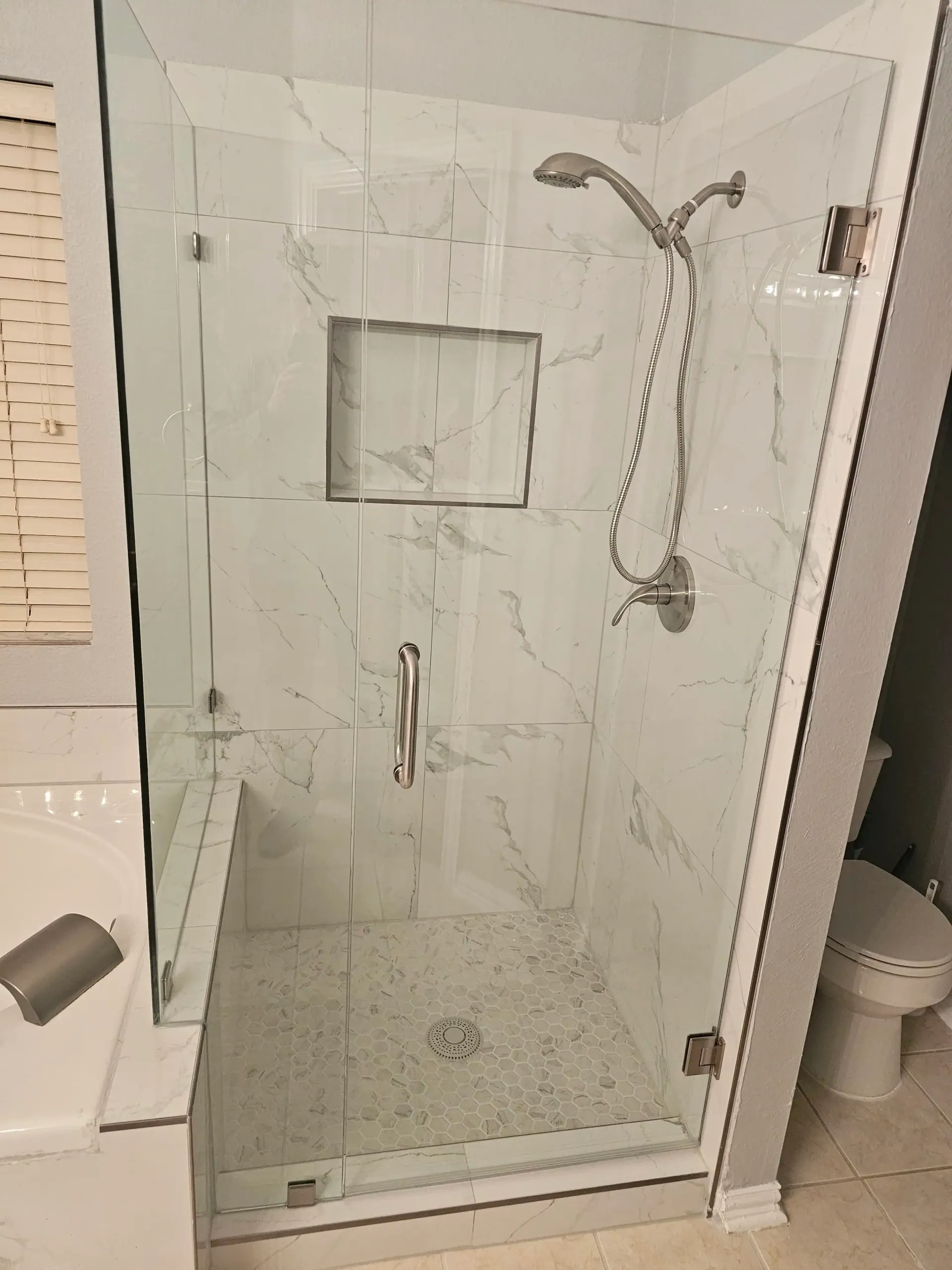 Glass shower enclosure with white marble-look tiles, a built-in shelf, and a shower head.