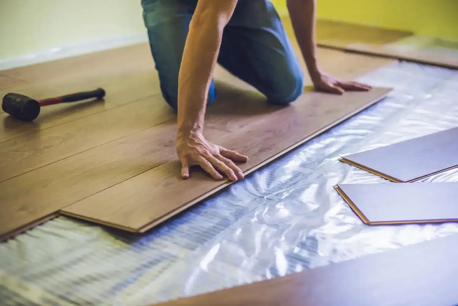 Person installing laminate flooring, kneeling, with hammer nearby.
