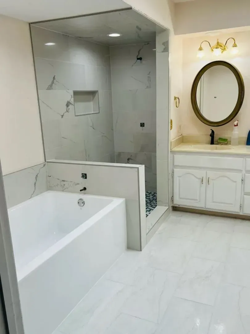 Modern bathroom with white tub, shower, and vanity. Marble-look tiles, gold mirror frame, and sconces.