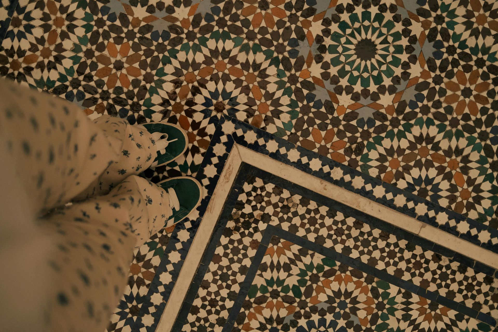 Feet in sneakers stand on a mosaic tiled floor with geometric patterns, with darker border tiles.