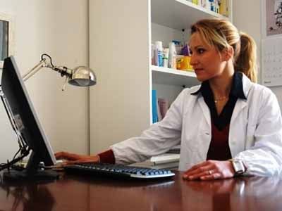 Una donna in camice da laboratorio è seduta a una scrivania e sta scrivendo al computer in quella che sembra una farmacia o uno studio medico.