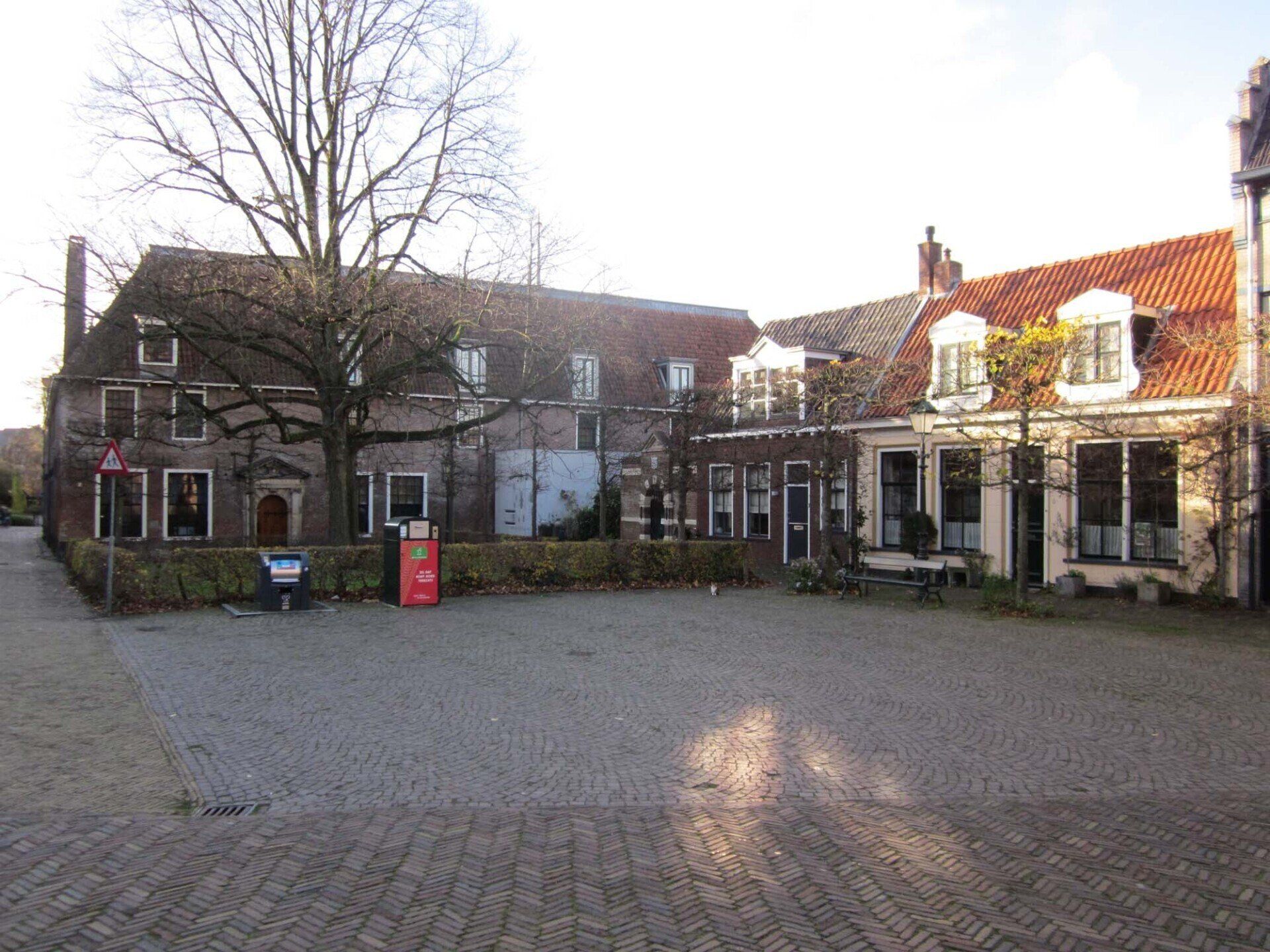 Rustige, met kinderkopjes bestrate binnenplaats met oude bakstenen gebouwen en een rood pannendak, badend in het zonlicht.
