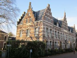 Historisch bakstenen gebouw met sierlijke gevels en hoge ramen, gelegen naast een met hagen omzoomde straat, badend in het daglicht.