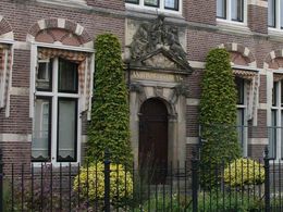 Een bakstenen gevel met hoge ramen, een sierlijke deuropening, klimop en een zwart ijzeren hek.