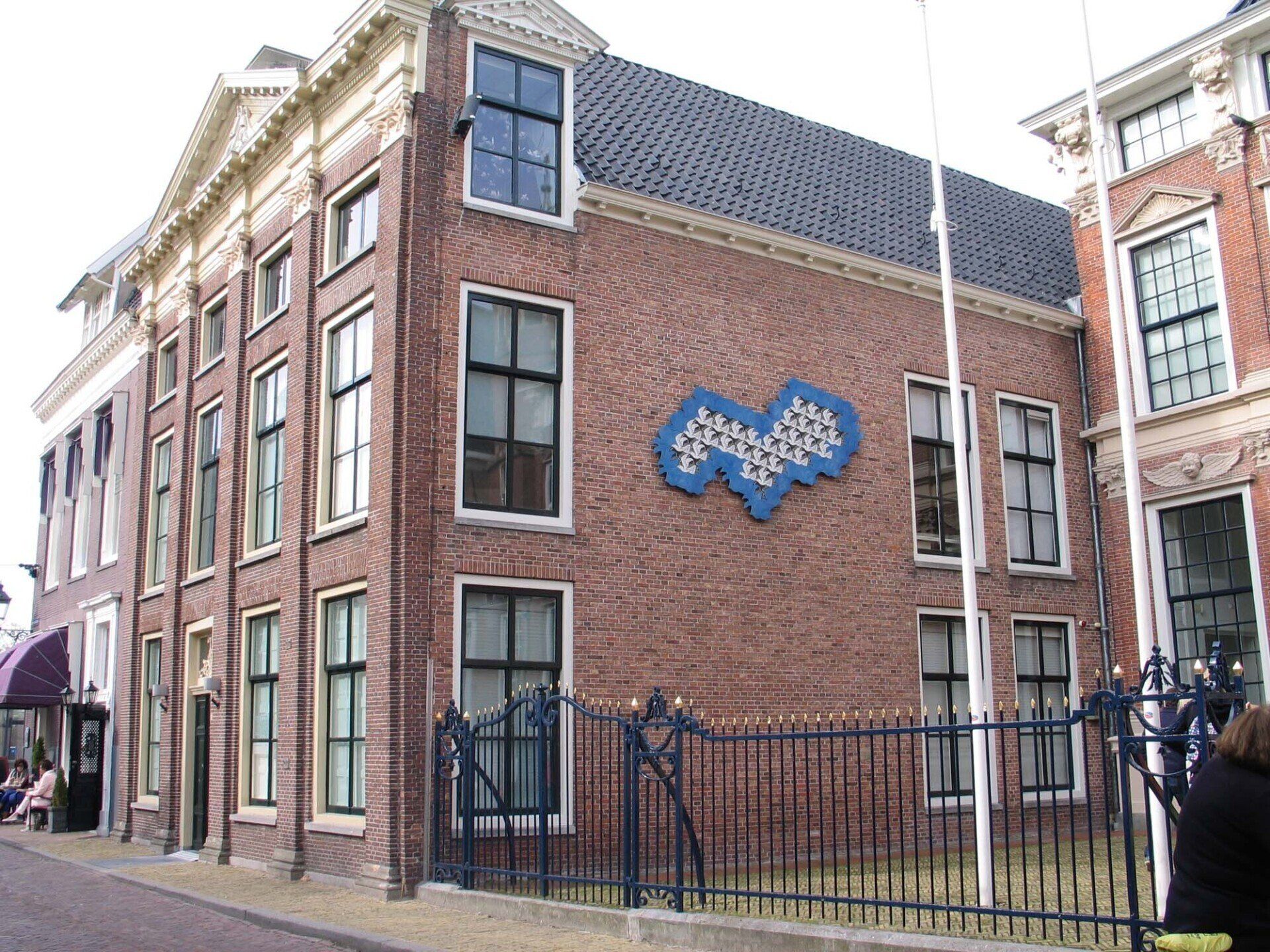 Bakstenen gebouw met blauwe decoratieve muurschilderingen en een zwart ijzeren hekwerk aan een stadsstraat.