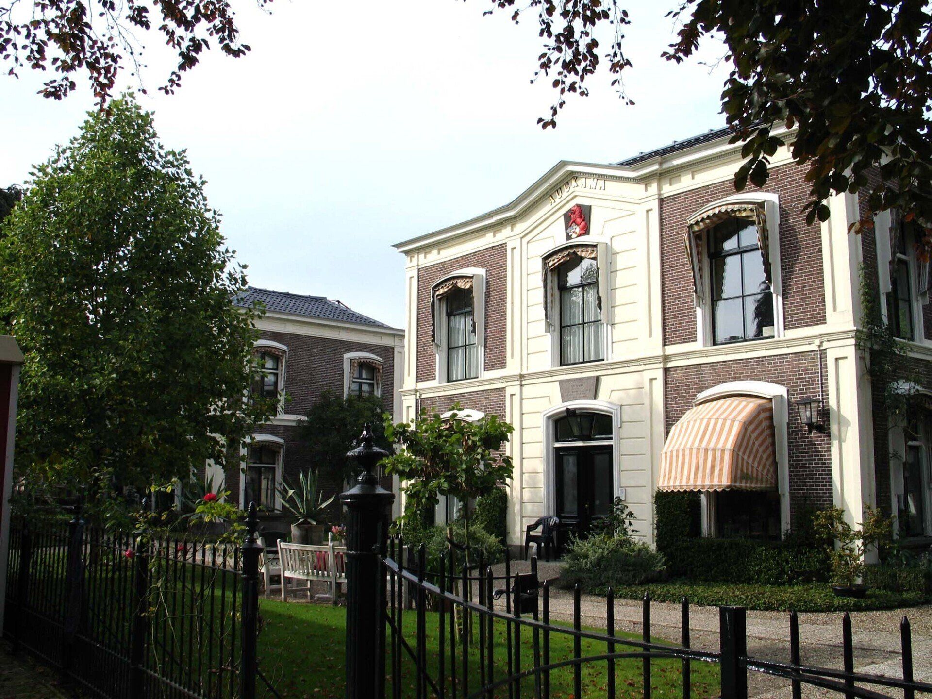 Elegant crèmekleurig herenhuis met rode accenten, boogvensters en een gestreepte luifel achter een zwart ijzeren hek.