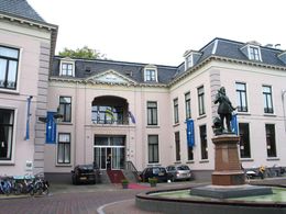 De binnenplaatsingang van een lichtroze gebouw met een fontein met een standbeeld ervoor en geparkeerde auto's.