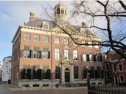 Historisch bakstenen gebouw met zwarte luiken en een kleine klokkentoren onder een kale boom.