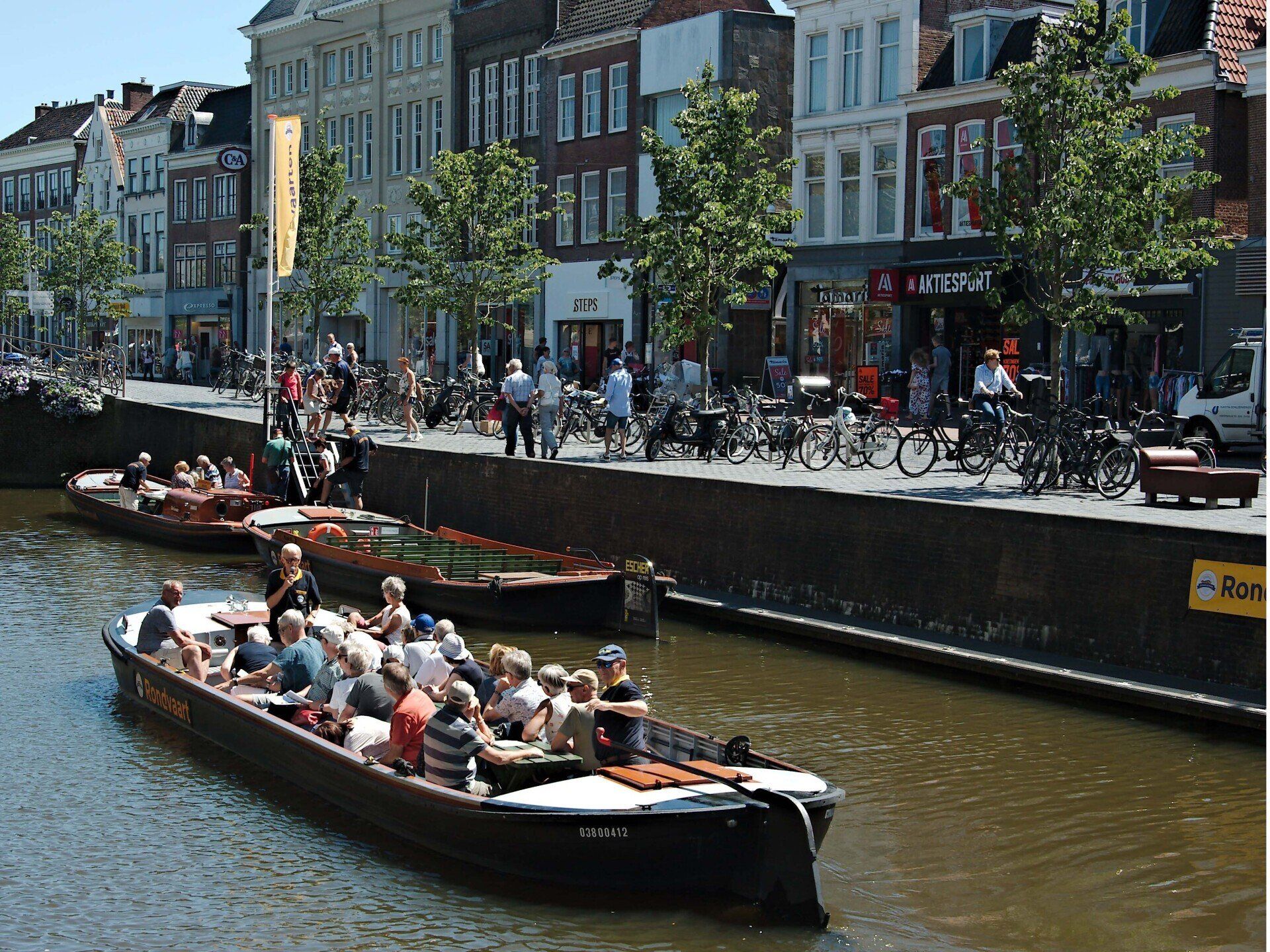 Rondvaartboten vol passagiers varen over een stadskanaal langs drukke gebouwen en fietsers aan de oever.
